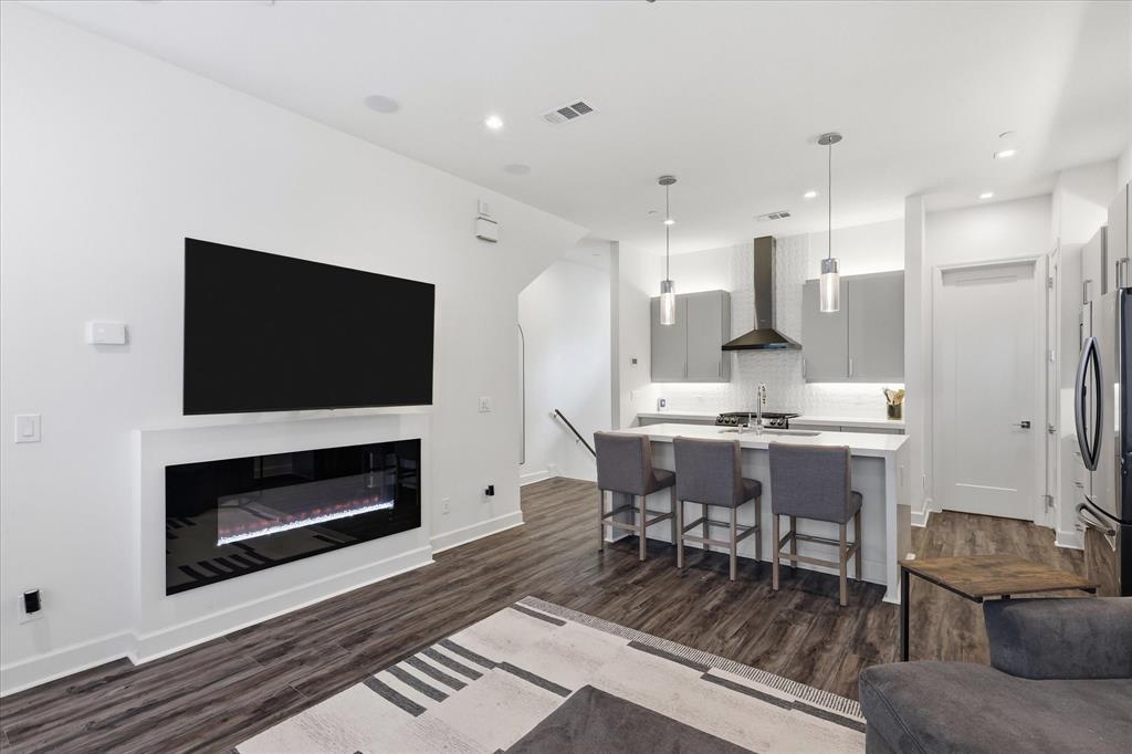 Linear wall-mounted electric fireplace, wood-finish flooring, kitchen island with stone countertop, stainless steel range hood, and pendant lighting
