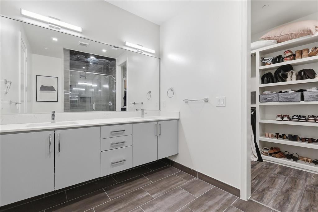 Floating dual-sink vanity with light gray cabinetry and white countertop, full-width wall mirror, wood-finish flooring, and walk-in closet featuring custom built-in shelving