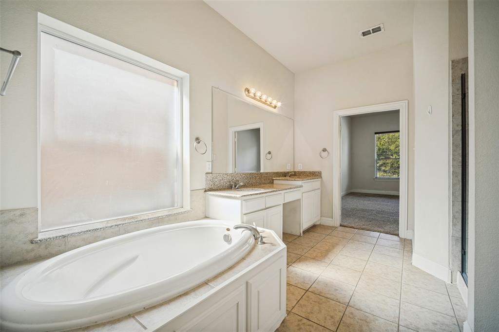 Bathroom featuring a drop-in soaking tub with a tiled surround, dual-sink vanity with stone-style countertops, expansive wall-mounted mirror, and tile flooring