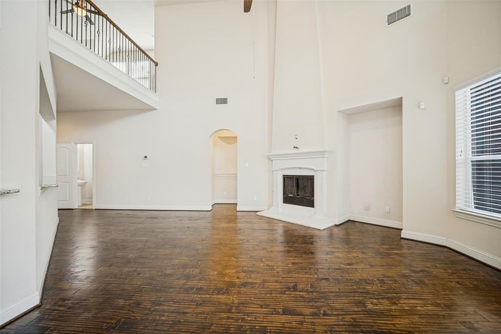 Double-height ceiling, fireplace with detailed mantel, wood-finish flooring, and an elevated mezzanine with a wrought iron railing