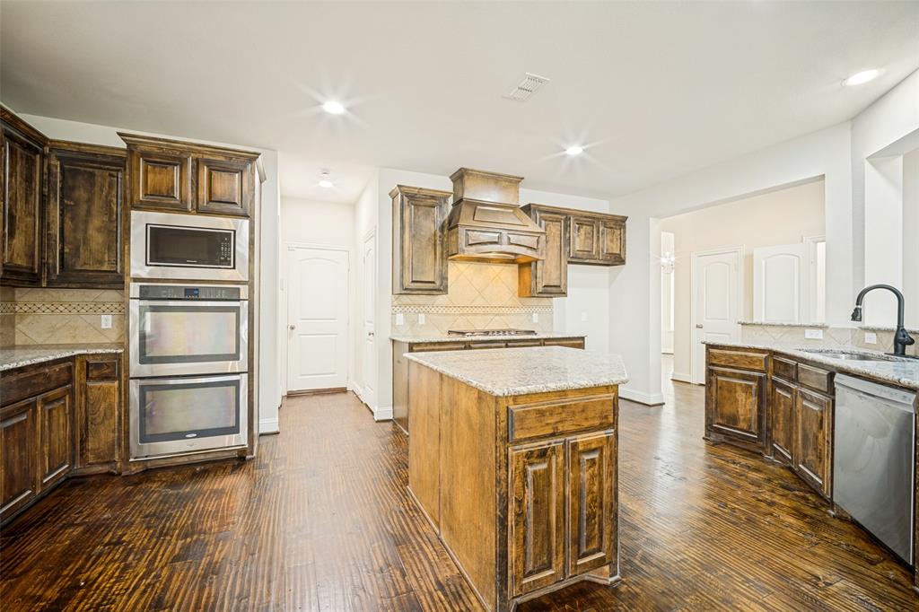 Kitchen featuring stained wood cabinetry, granite countertops, integrated double wall ovens, built-in range hood, and wood-finish flooring Kitchen featuring stained wood cabinetry, granite countertops, integrated double wall ovens, built-in range hood, and wood-finish flooring