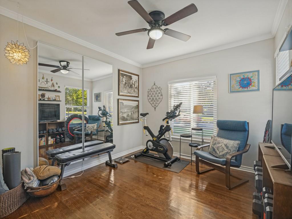 Room featuring wood-finish flooring, full-wall mirrored paneling, a ceiling fan with integrated lighting, crown molding, and horizontal slat window blinds