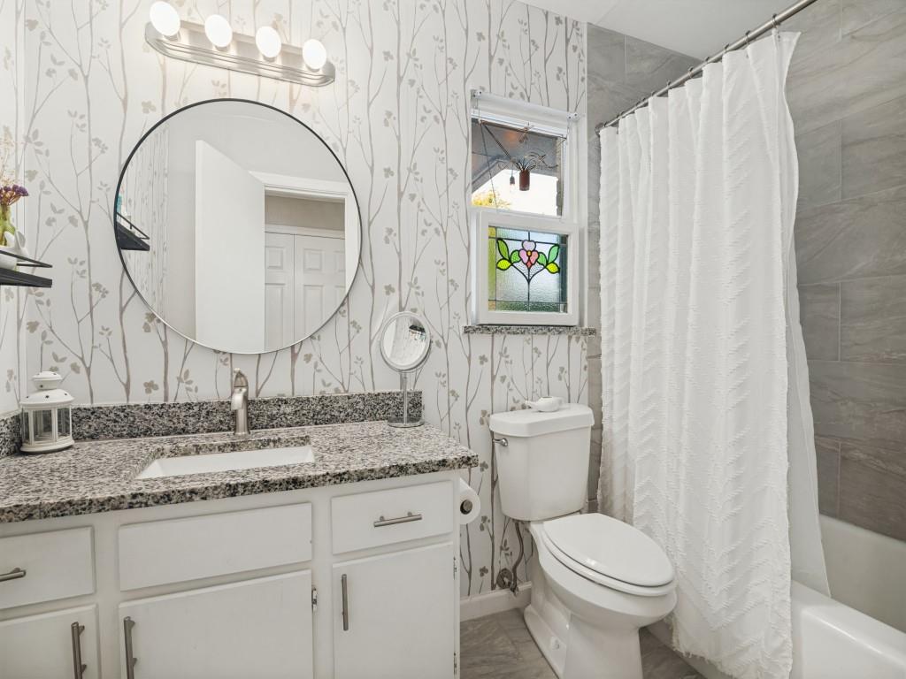 Bathroom featuring a vanity with stone-finish countertop, round mirror, patterned wall covering, window with decorative stained-glass inlay, and shower-tub combination with grey tile surround