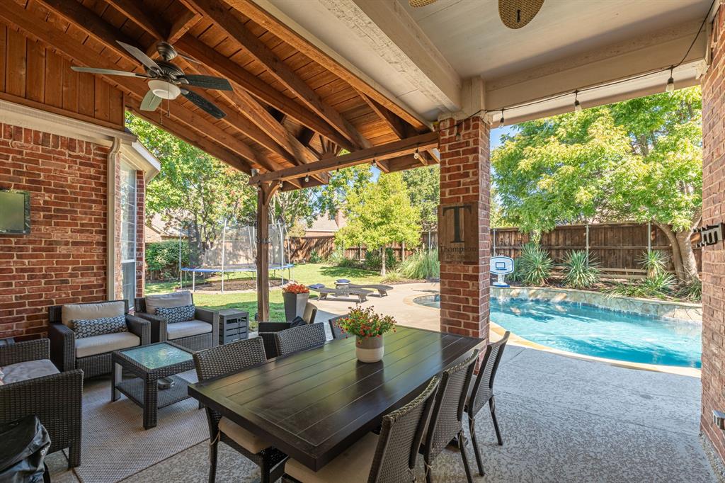 Covered patio featuring a wood-plank ceiling, exposed timber beams, brick columns, a concrete floor, and an integrated ceiling fan, overlooking a swimming pool and landscaped grounds