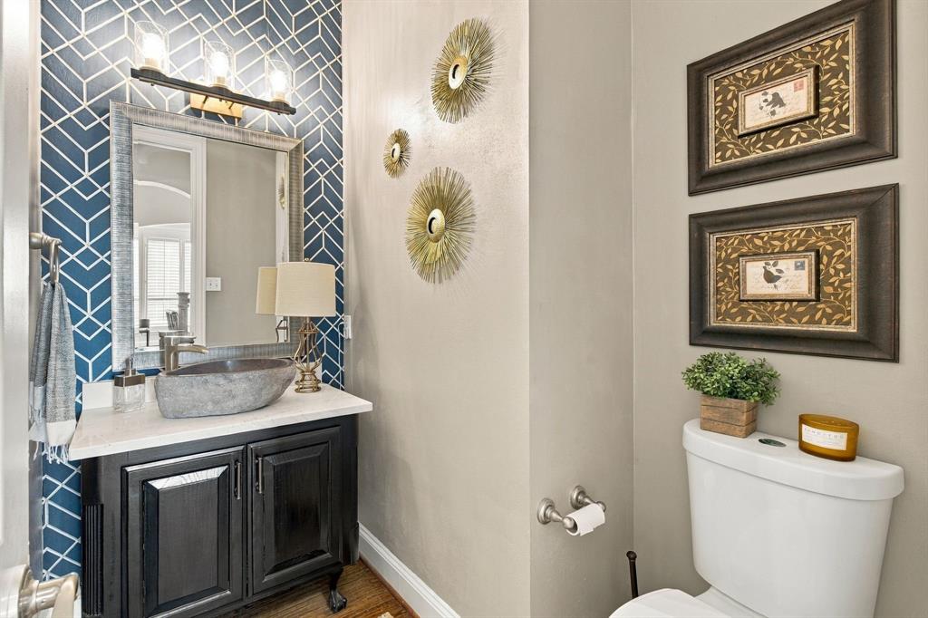 Black vanity cabinet with a light-toned countertop, stone-finish vessel sink, geometric patterned accent wall, and three-light vanity fixture