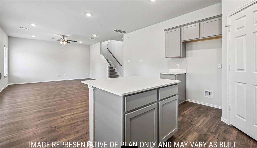 Kitchen island with white countertop and grey cabinetry, wood-finish flooring, recessed lighting, and a staircase featuring a wooden handrail Kitchen island with white countertop and grey cabinetry, wood-finish flooring, recessed lighting, and a staircase featuring a wooden handrail