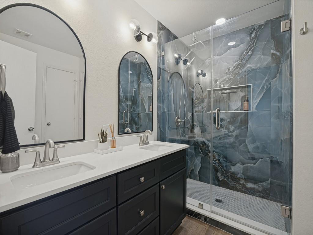 Dual vanity with solid-surface countertop and dark cabinetry, arched wall mirrors, and walk-in glass-enclosed shower featuring large-format patterned wall tiles and a built-in recessed niche