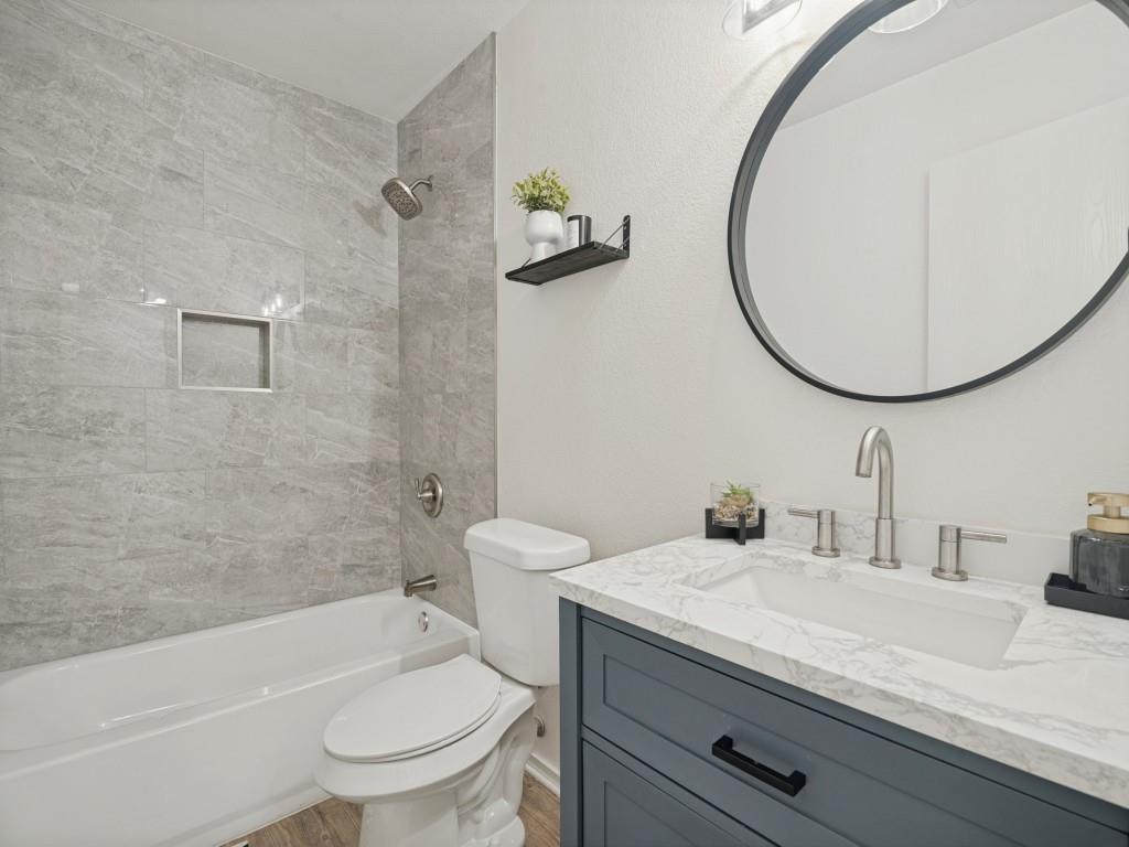 Bathroom featuring a vanity with shaker-style cabinetry, stone-finish countertop, integrated rectangular sink, wall-mounted circular mirror, and tub-shower combination with grey tile surround and built-in recessed niche