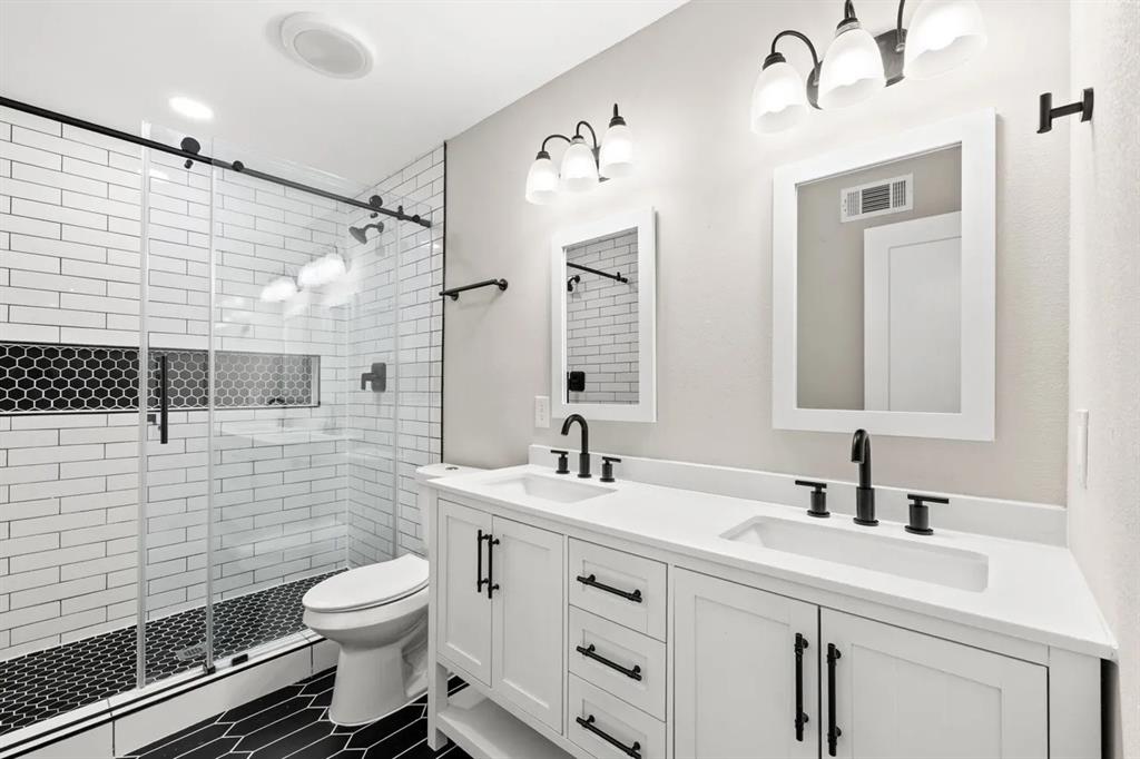 Dual-sink vanity with a white solid-surface countertop and matte black hardware, walk-in shower featuring a glass enclosure with sliding track, white subway wall tile, and hexagonal black floor tile