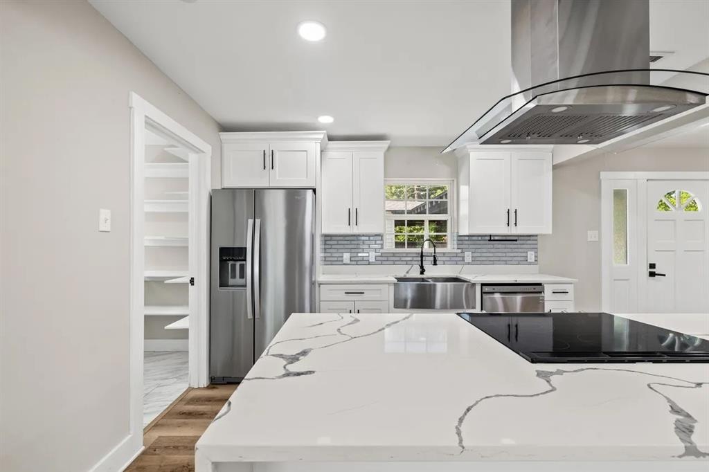 Kitchen featuring an oversized stone-finish waterfall island, stainless steel range hood, farmhouse sink, shaker-style cabinetry, and a walk-in pantry