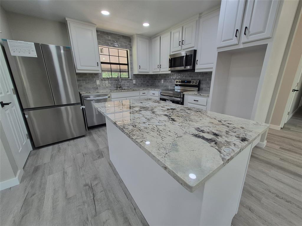Kitchen featuring a central island with stone countertop, white shaker-style cabinetry, stainless steel appliances, decorative tile backsplash, and wood-finish flooring
