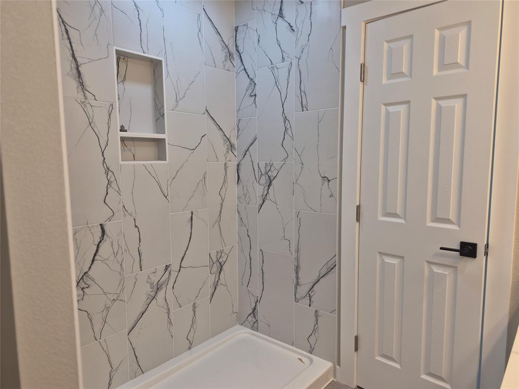 Shower enclosure featuring floor-to-ceiling large-format tile with decorative veining, built-in recessed storage niche, white acrylic shower basin, and a six-panel door with matte black hardware