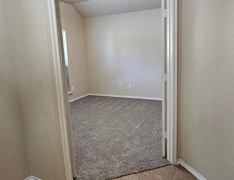 Neutral-toned interior space featuring textured wall-to-wall carpeting, vaulted ceiling, dual-swing doors, and a window with fitted blinds