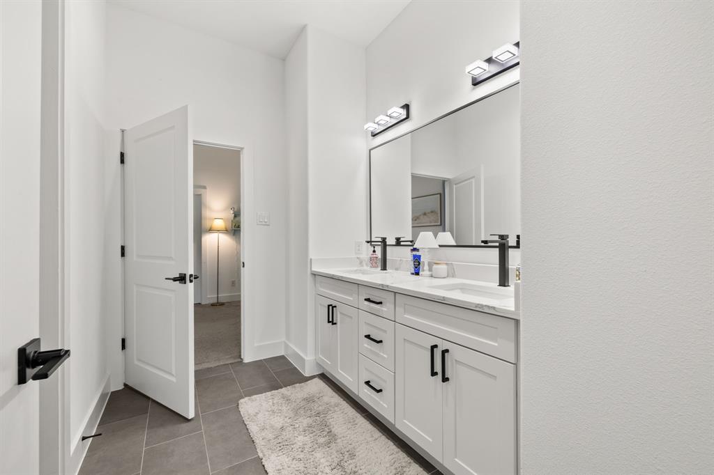 Dual-sink vanity featuring white cabinetry, stone-finish countertops, matte black hardware, and wall-mounted matte black faucets, complemented by two modern light fixtures and a full-width mirror