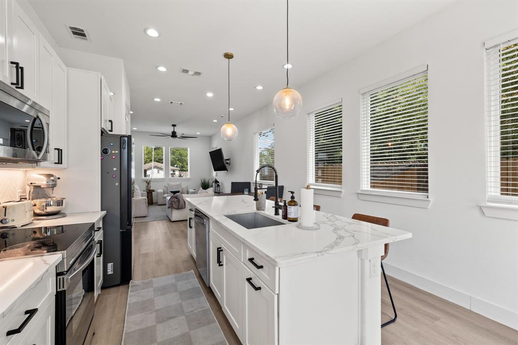 Kitchen featuring a central island with a stone-finish countertop, white cabinetry with matte black hardware, stainless steel appliances, wood-finish flooring, and pendant lighting Kitchen featuring a central island with a stone-finish countertop, white cabinetry with matte black hardware, stainless steel appliances, wood-finish flooring, and pendant lighting