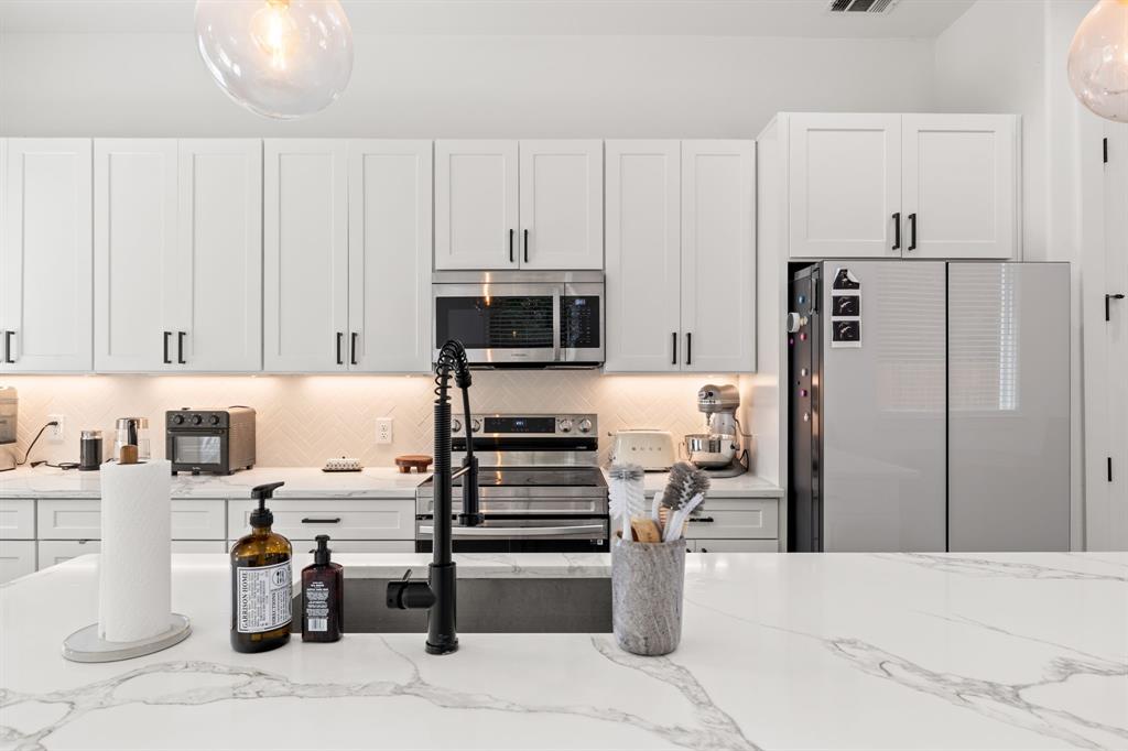 Kitchen featuring white Shaker-style cabinetry, a large island with a stone-surface countertop, herringbone-patterned backsplash, stainless steel appliances, and a matte black gooseneck faucet Kitchen featuring white Shaker-style cabinetry, a large island with a stone-surface countertop, herringbone-patterned backsplash, stainless steel appliances, and a matte black gooseneck faucet
