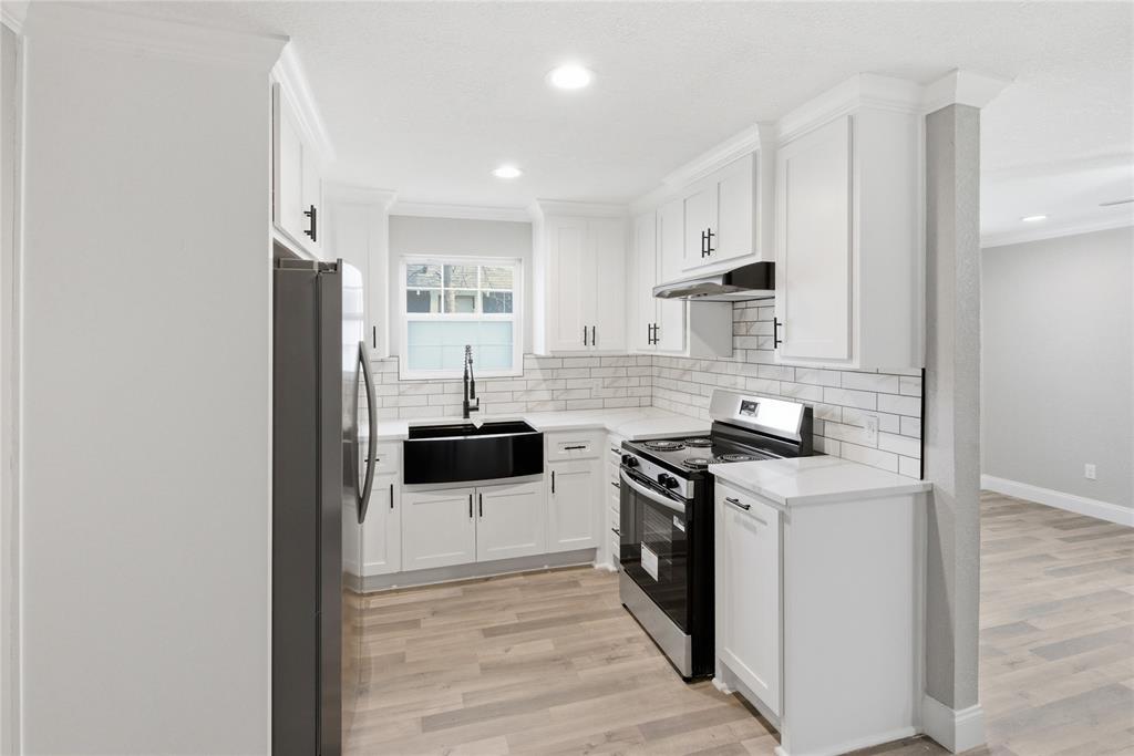 Kitchen featuring white shaker-style cabinetry, light-toned countertop surfaces, white subway tile backsplash, farmhouse-style apron sink with a high-arch faucet, and wood-finish flooring Kitchen featuring white shaker-style cabinetry, light-toned countertop surfaces, white subway tile backsplash, farmhouse-style apron sink with a high-arch faucet, and wood-finish flooring