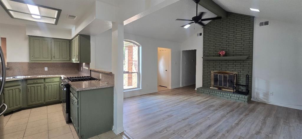 Kitchen featuring cabinetry, a gas cooktop, and stone countertops, paired with a living area showcasing wood-finish flooring, a vaulted ceiling, and a full-height brick fireplace