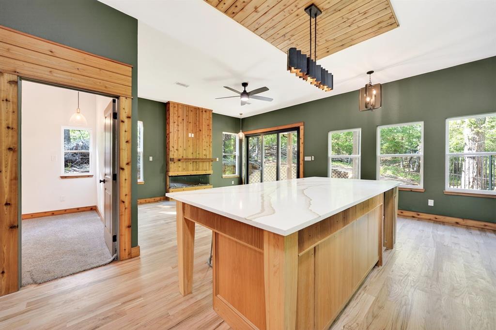 Kitchen featuring an expansive island with a light-toned stone countertop, wood-finish flooring, a wood-paneled ceiling accent, a floor-to-ceiling wood fireplace surround, and multiple large windows