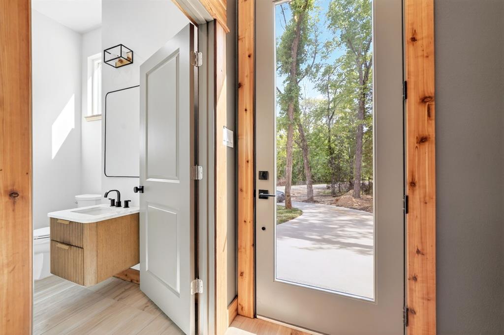 Modern half-bath featuring a floating wood-finish vanity, rectangular vessel sink, matte black faucet, and geometric wall-mounted light fixture, complemented by an entry door with natural wood casing