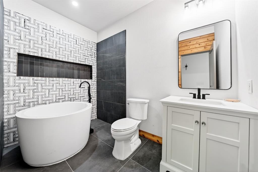 Freestanding soaking tub with matte black wall-mounted faucet, geometric tiled accent wall featuring a recessed niche, white vanity with rectangular sink, and large-format dark floor tile