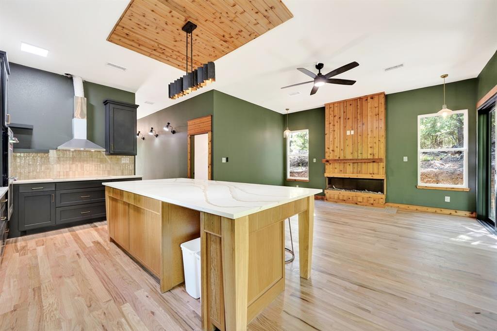 Kitchen featuring an oversized island with a quartz countertop, wood-finish flooring, wood-paneled fireplace surround, integrated ceiling-mounted wood feature, and a modern linear pendant light fixture