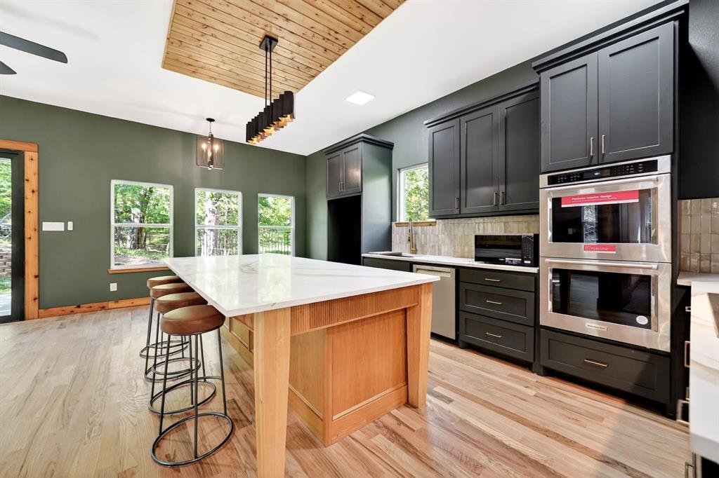 Kitchen featuring a natural-finish wood center island with a stone countertop, dark cabinetry, stainless steel double wall ovens, wood-finish flooring, and a recessed ceiling accent with plank wood detailing Kitchen featuring a natural-finish wood center island with a stone countertop, dark cabinetry, stainless steel double wall ovens, wood-finish flooring, and a recessed ceiling accent with plank wood detailing