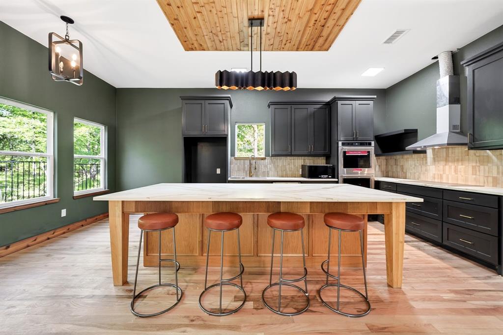 Kitchen featuring a central island with a stone countertop, wood-finish cabinetry, built-in double oven, stainless steel range hood, and a ceiling inset with wood-plank detailing Kitchen featuring a central island with a stone countertop, wood-finish cabinetry, built-in double oven, stainless steel range hood, and a ceiling inset with wood-plank detailing