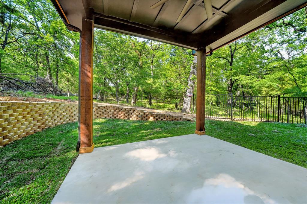 Covered patio featuring concrete flooring and wooden support columns, complemented by a tiered stone retaining wall and black metal fencing