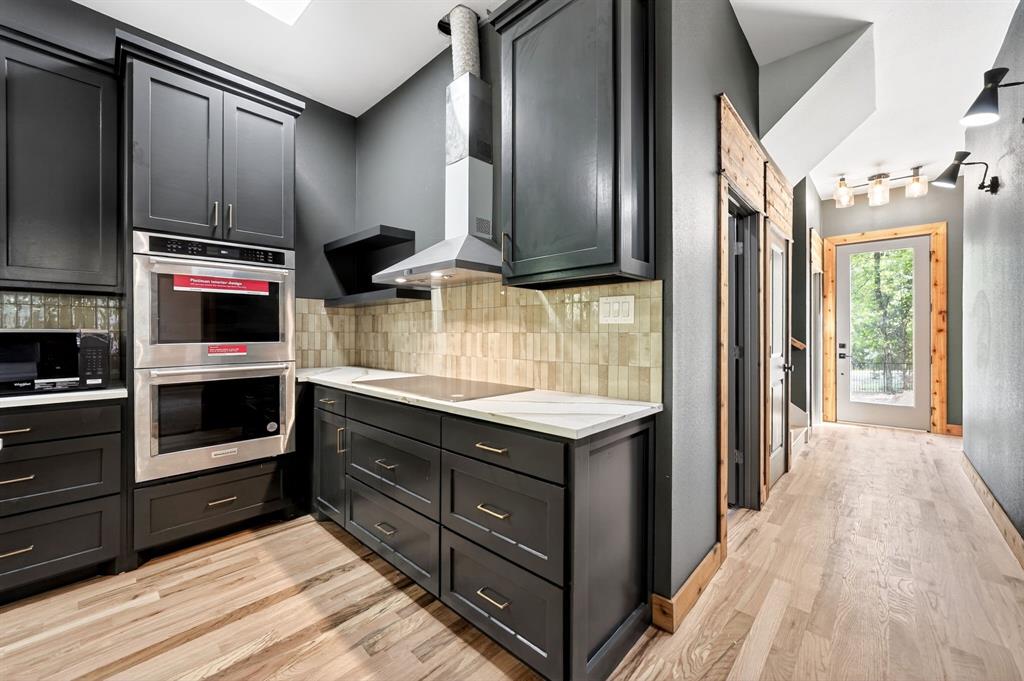 Custom dark-finish cabinetry featuring brass-tone hardware, stainless steel built-in double ovens, natural stone-finish countertops, textured tile backsplash, and light wood-finish flooring
