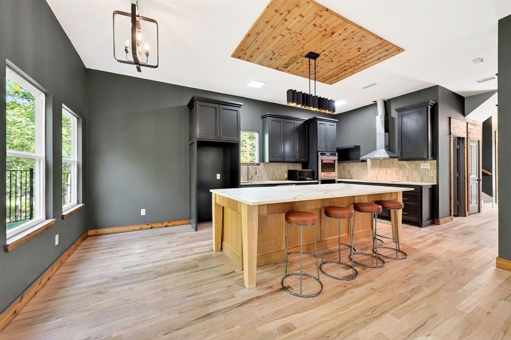 Kitchen featuring an oversized island with a stone-top surface, wood-finish flooring, custom cabinetry with dark finish, a wood-paneled ceiling inset, and stainless steel hood range Kitchen featuring an oversized island with a stone-top surface, wood-finish flooring, custom cabinetry with dark finish, a wood-paneled ceiling inset, and stainless steel hood range