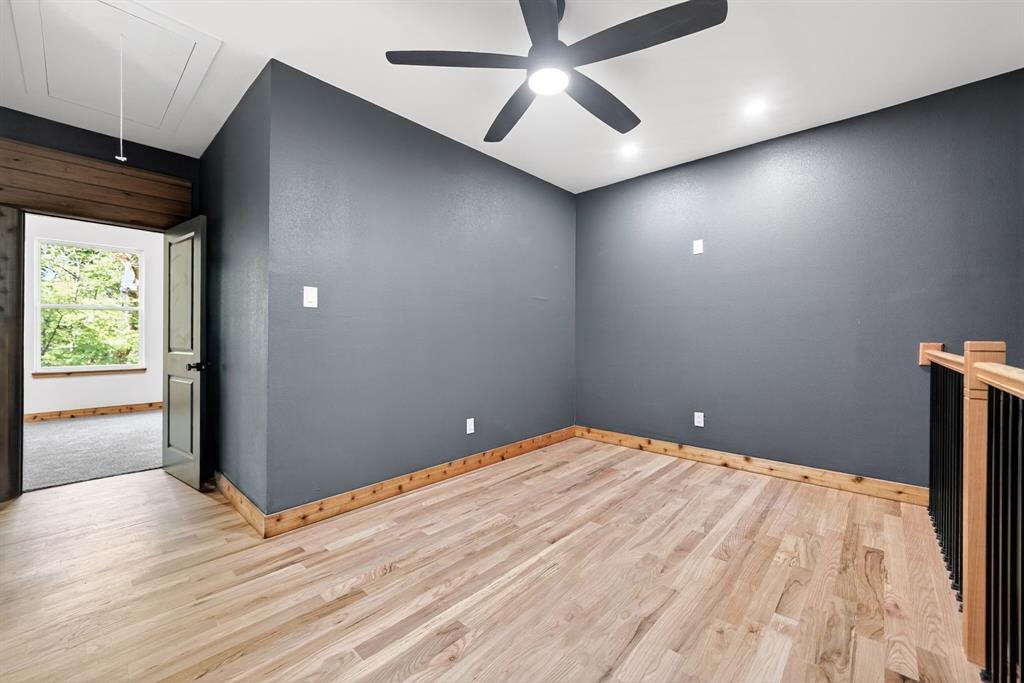 Wood-finish flooring, dark-toned interior walls, natural wood trim, matte black ceiling fan, and staircase with wooden handrail and black metal balusters