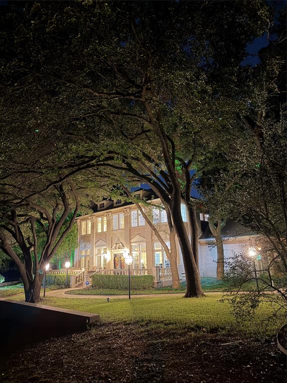 Two-story brick exterior featuring a stone balustrade, mature canopy trees, manicured landscaping, and integrated exterior lighting