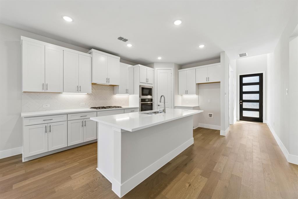 Kitchen featuring white cabinetry, quartz countertops, wood-finish flooring, a herringbone-patterned tile backsplash, and a central island with a stainless steel sink