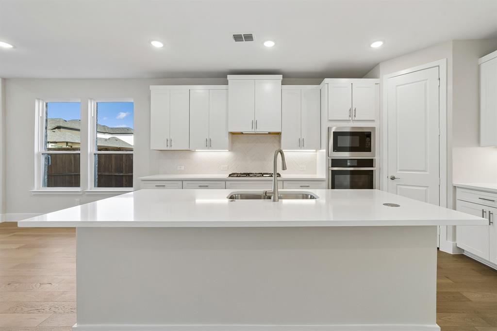 Kitchen featuring a large central island with a solid surface countertop, white shaker-style cabinetry, stainless steel built-in double oven, gas cooktop, and wood-finish flooring