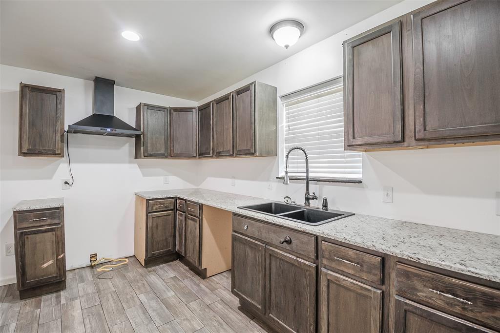 Kitchen featuring stained wood cabinetry, light-toned stone-finish countertops, dual-basin under-mount sink with a high-arch matte black faucet, wall-mounted range hood, and wood-finish flooring
