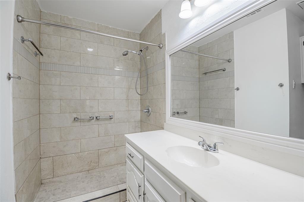 Bathroom featuring a walk-in shower with stone-finish tile surround, an integrated handheld shower head, a white cabinetry vanity with a solid-surface countertop, and a large-format vanity mirror with trim detail