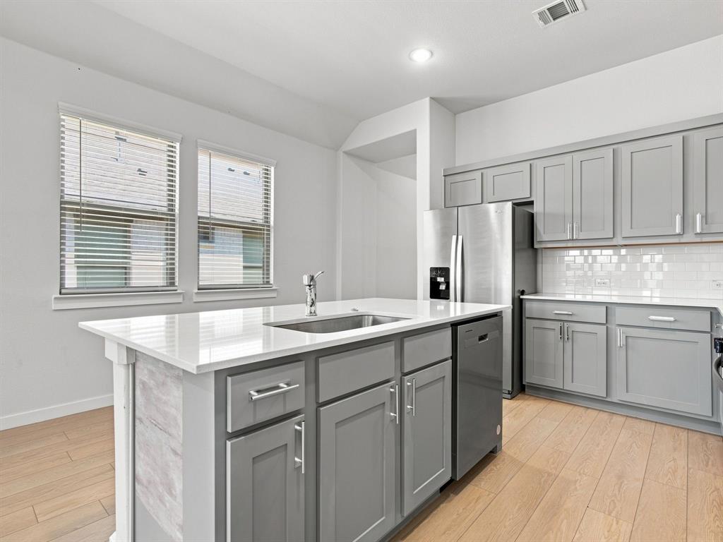 Kitchen featuring grey cabinetry, white countertops, a central island with a stainless steel undermount sink, subway tile backsplash, and light-toned wood-finish flooring