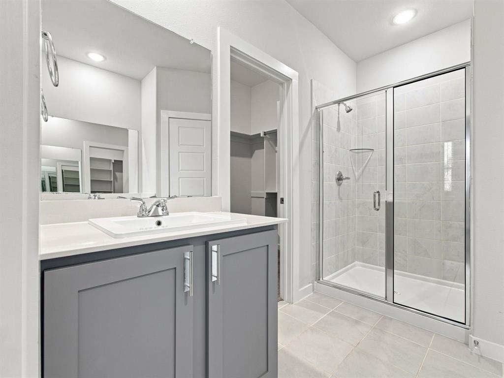Grey cabinetry with silver-tone hardware, rectangular undermount sink, quartz countertop, large wall-mounted mirror, and glass-enclosed shower with tiled surround