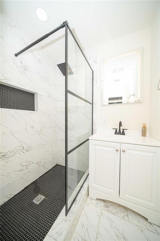 Walk-in shower with matte black-framed glass enclosure, stone-patterned wall tile, recessed tiled niche, and dark penny-round flooring, complemented by a white vanity with a top-mounted faucet and square mirror