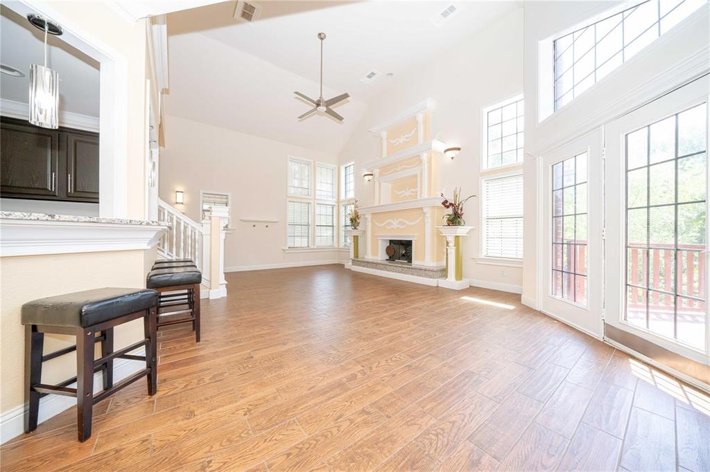 Open-concept living space featuring vaulted ceilings, wood-finish flooring, a decorative fireplace mantle with stone-surround hearth, and double glass-paneled exterior doors with an arched transom window Open-concept living space featuring vaulted ceilings, wood-finish flooring, a decorative fireplace mantle with stone-surround hearth, and double glass-paneled exterior doors with an arched transom window