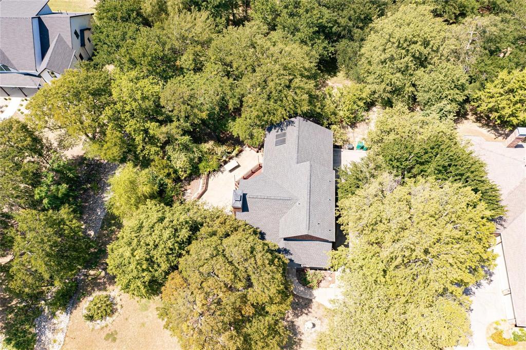 Aerial perspective featuring a multi-pitched asphalt shingle roof, solar panels, stone hardscaping, and mature trees on a wooded lot