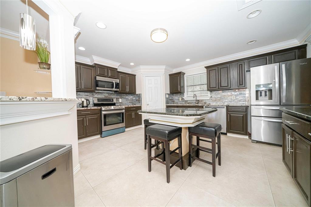 Kitchen featuring dark wood cabinetry, a central island with a stone countertop, stone-tiled backsplash, stainless steel appliances, and neutral tile flooring Kitchen featuring dark wood cabinetry, a central island with a stone countertop, stone-tiled backsplash, stainless steel appliances, and neutral tile flooring