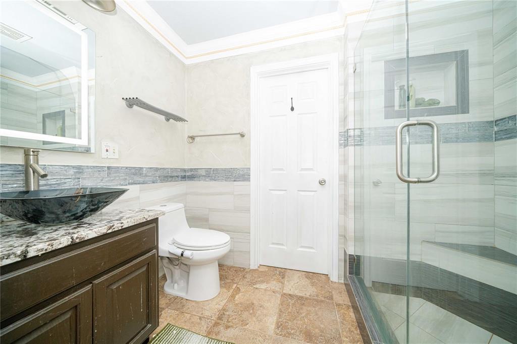 Bathroom featuring dark wood-finish cabinetry, patterned stone-surface countertop, glass vessel sink, and glass-enclosed shower with tiled walls and built-in shelving