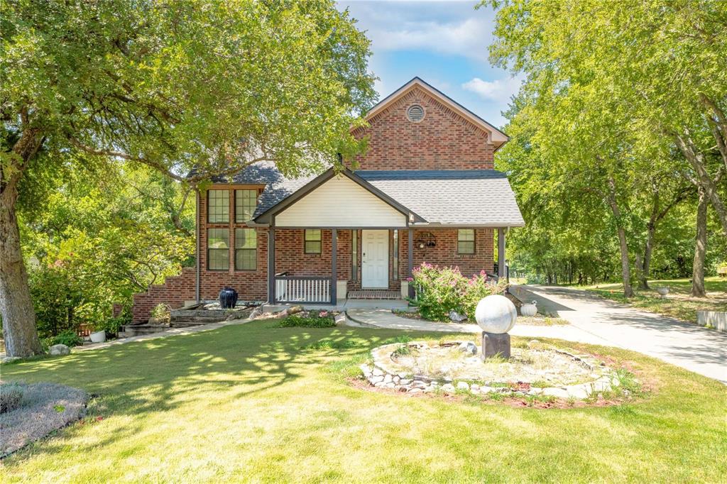 Multi-story brick exterior with a gabled roofline, covered front porch, stone-lined landscaping, and mature trees Multi-story brick exterior with a gabled roofline, covered front porch, stone-lined landscaping, and mature trees