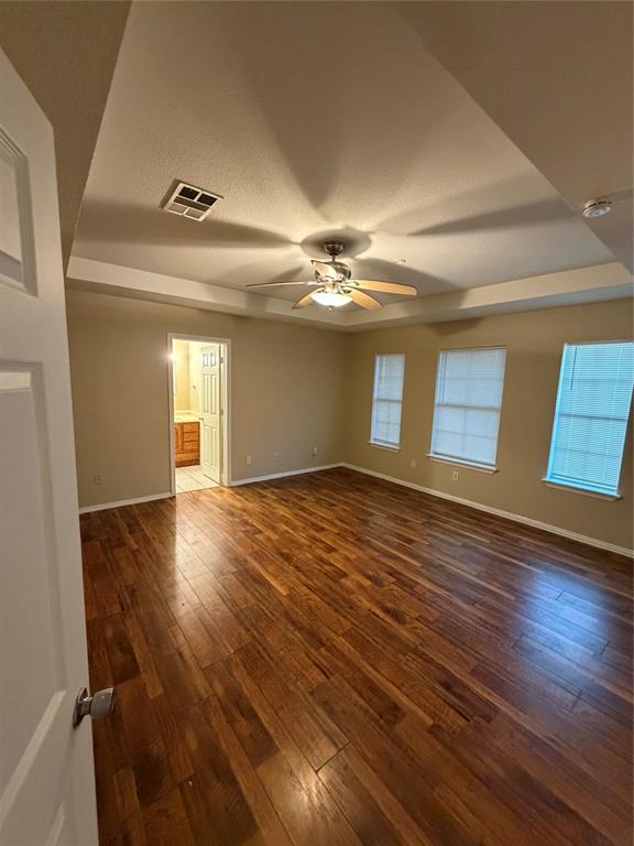 Room featuring wood-finish flooring, tray ceiling with a central multi-blade ceiling fan, and three windows Room featuring wood-finish flooring, tray ceiling with a central multi-blade ceiling fan, and three windows