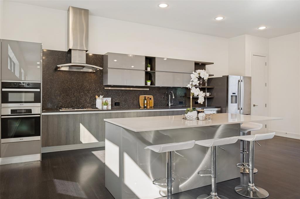 Modern kitchen featuring flat-panel cabinetry, a central island with a quartz countertop, stainless steel appliances including a built-in double oven and range hood, and dark-toned wood-finish flooring