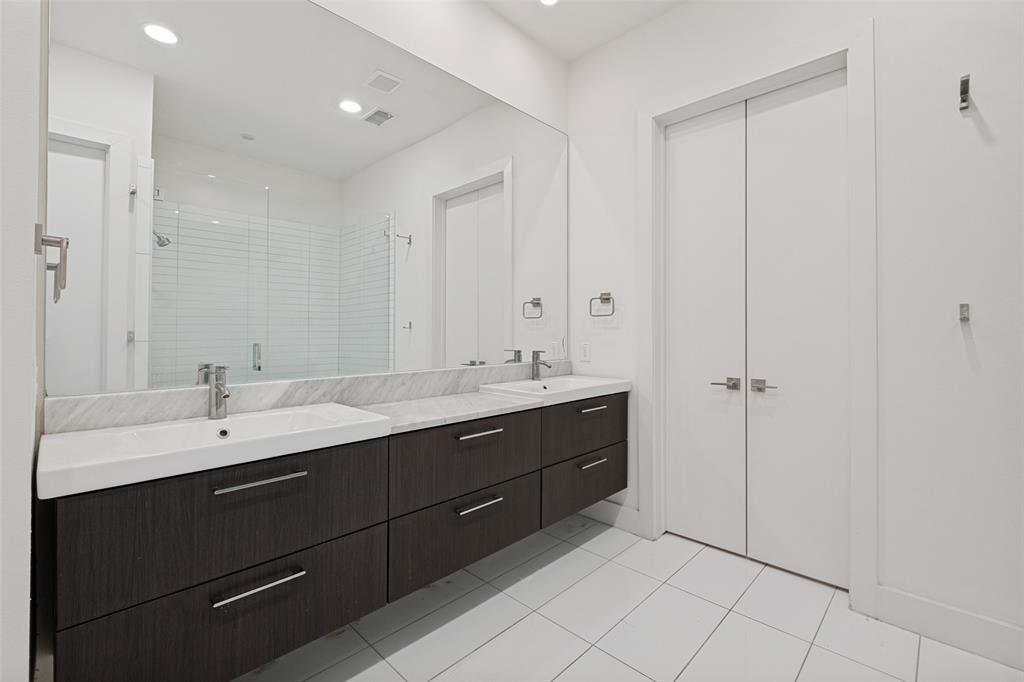 Floating double vanity with dark wood-finish cabinetry, stone countertop, integrated sinks, and expansive wall-to-wall mirror, complemented by floor-to-ceiling white tile flooring