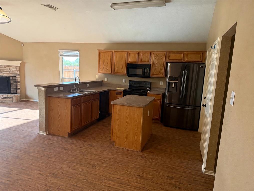 Kitchen featuring wood cabinetry, an island with countertop surface, wood-finish flooring, and a brick fireplace Kitchen featuring wood cabinetry, an island with countertop surface, wood-finish flooring, and a brick fireplace