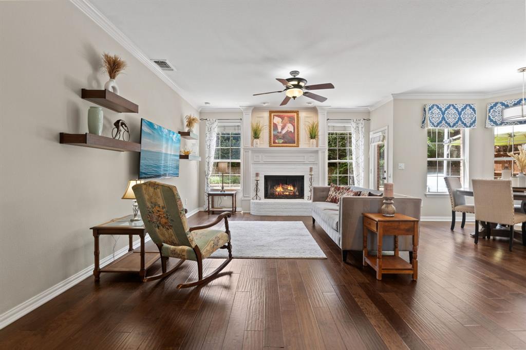 Open-concept living space featuring wood-finish flooring, a white-mantel fireplace with ornate columns, crown molding, a ceiling fan with integrated lighting, and multiple windows with deep sills Open-concept living space featuring wood-finish flooring, a white-mantel fireplace with ornate columns, crown molding, a ceiling fan with integrated lighting, and multiple windows with deep sills